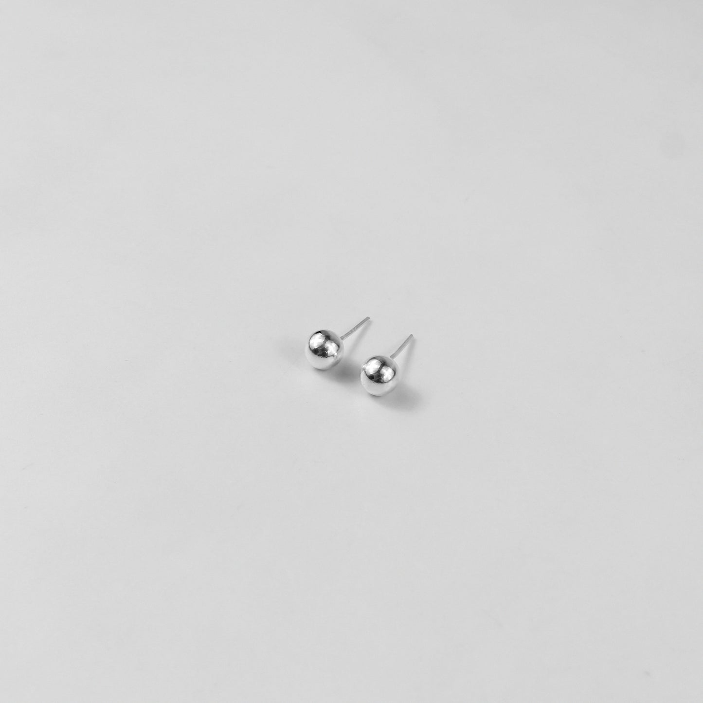Sterling silver ear studs shaped like perfect spheres, displayed on a white marble background. Their polished texture creates a luxurious and timeless appeal.